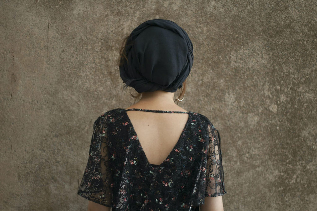 A person faces a cement wall. They are wearing a black gauzy, floral shirt and hoop earrings. Wisps of brown hair escape the black head scarf they are wearing.