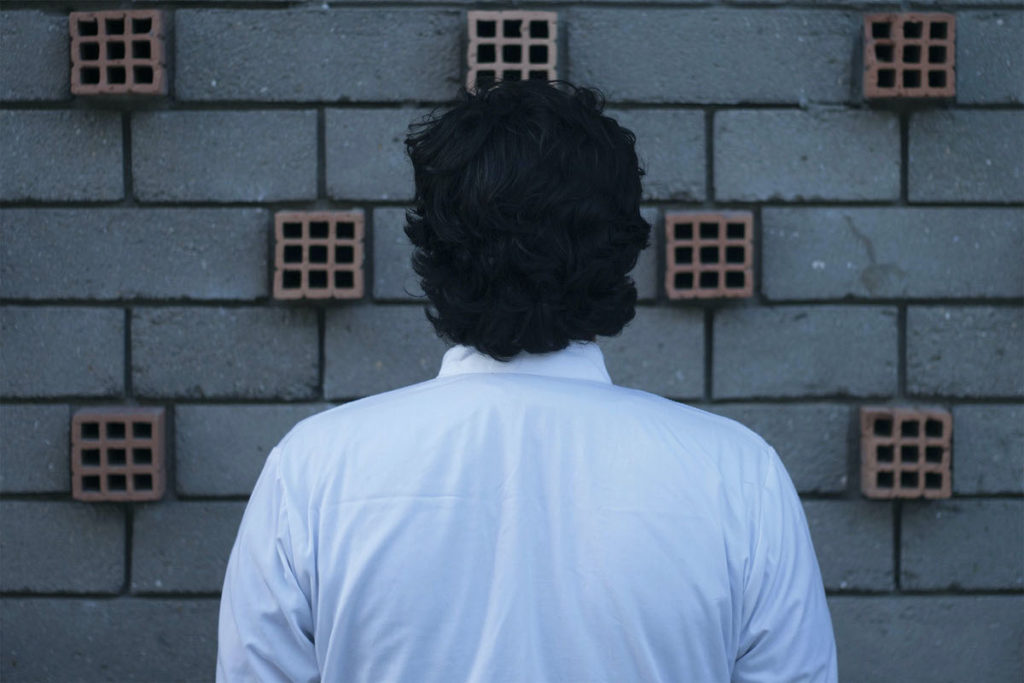 A person with short, wavy dark hair wears a white long-sleeved shirt and stands in front of a gray brick wall. Red, square bricks are sporadically interspersed between the gray bricks.