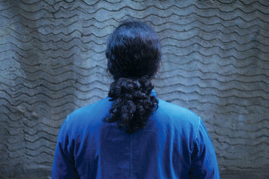 A person with dark, curly hair pulled into a ponytail wears a long-sleeved blue shirt. They face a cement wall with an uneven, wavy pattern carved into it.