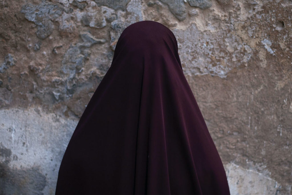 A person in a maroon hijab faces a crumbling cement wall.