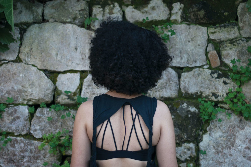 A person with dark, natural hair faces a stone wall with weeds crowing between uneven cracks. They are wearing a black tank top with an exposed back.