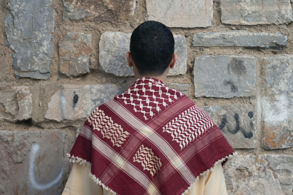 Photo of a person facing a stone wall with hints of spray paint on it. The person has close-cropped dark hair and a red-and-white scarf is draped over their shoulders.