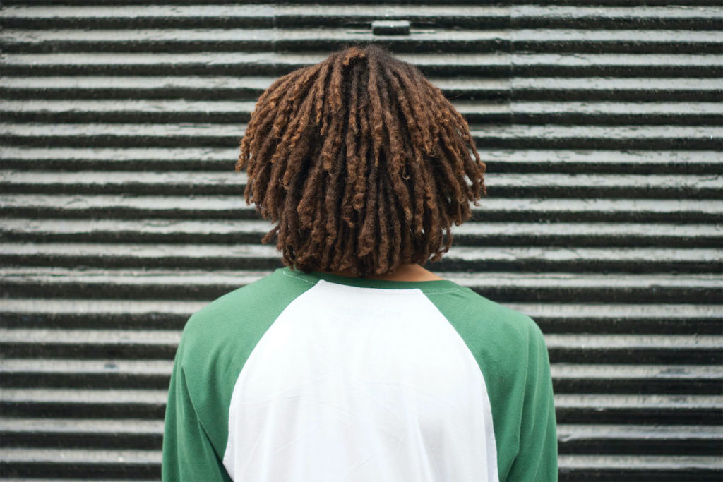Photo of a person with short dreadlocks in a green-and-white baseball shirt facing a ridged gray wall.